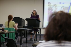 A woman is speaking to people in front of a projector screen.