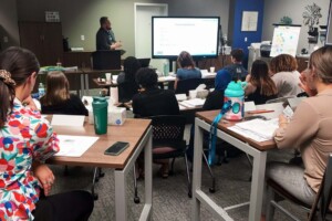 A group of people sitting at tables in front of a projector screen.